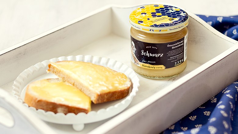 Honig, © Sooo gut schmeckt die Bucklige Welt/ Viktoria Kornfeld Ein weißes Holztablett mit einem Glas Honig und einem weißen Teller mit zwei Scheiben Honigbrot.