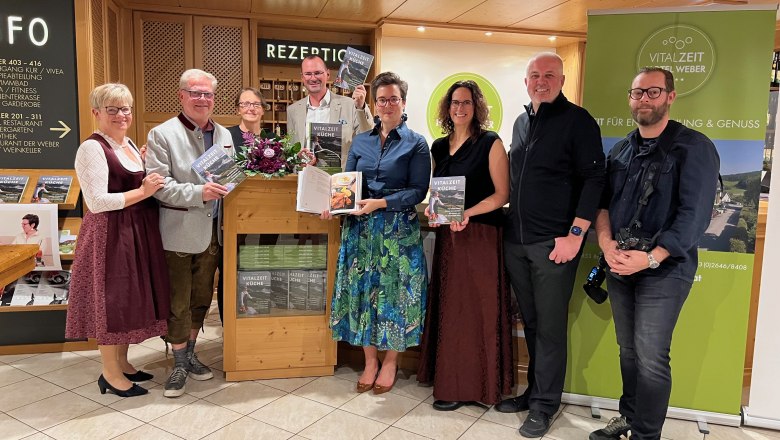 vlnr.: Petra Weber, Autor Alfred Weber, Grafikerin Anna Schiefer, Thomas Scherz, Mag. Katrin Scherz-Kogelbauer, Lektorin Elisabeth Zenz, Food-Fotograf Markus Proske, Fotograf Thomas Gobauer, © Scherz-Kogelbauer Mehrere Personen stehen vor der Rezeption im VitalZeit Hotel Weber, einige halten ein Kochbuch in der Hand.