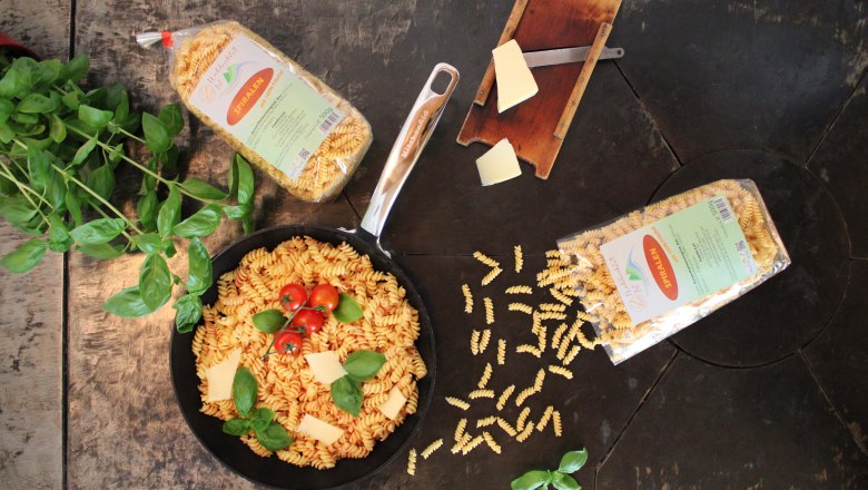 Teigwaren vom Feinsten, © Familie List Eine Pfanne mit Spiralnudeln, Tomaten und Basilikum auf einem Holztisch, umgeben von Nudelpackungen und einem Käsehobel.