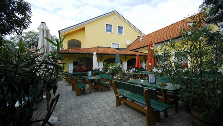Gastgarten Heuriger & Weingut Döller, © Familie Döller Gastgarten mit grünen Tischen und Bänken sowie geschlossenen Sonnenschirmen vor einem gelben Gebäude.