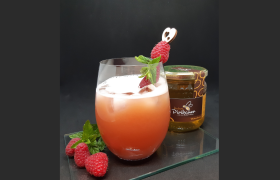 Honey Rider Cocktail, © Imkerei Piribauer Ein Glas mit Honey Rider Cocktail, Eiswürfeln und Himbeerspieß, daneben Himbeeren und Honigglas.