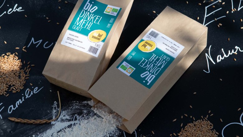 Bio-Dinkelmehl der Familie Trimmel, © Biohof Trimmel / Ulrike Korntheuer Zwei Packungen Bio-Dinkelmehl auf schwarzem Hintergrund mit Getreidekörnern und weißen Schriftzügen umgeben.