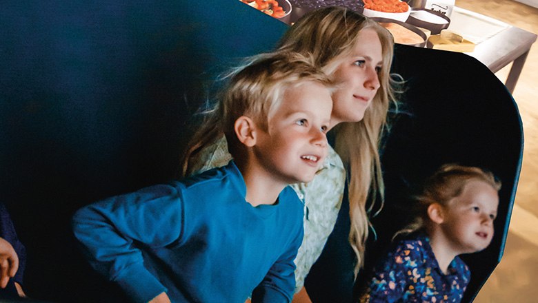 Eis-Greissler Eis-Zeitreise & Flying-Theater, © Blochberger Eisproduktion GmbH Zwei Kinder und eine Frau sitzen in einem Waggon im Flying-Theater und schauen gebannt nach vorne.