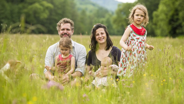 Die Familie, © Dreikanterei Fröhliche Familie mit drei Kindern sitzt auf einer Blumenwiese mit Bäumen im Hintergrund.