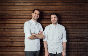 Die Brüder: Martin und Stefan Görg, © Wiener Alpen, Sophie Menegaldo Zwei Männer in weißen Hemden stehen vor einer Holzwand und lächeln.