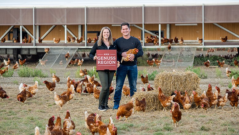 Daniela und Klaus Stocker, © Stocker Ei/Martina Siebenhandl Eine Frau mit einem roten Schild der "AMA Genusregion" und ein Mann mit einem Huhn stehen vor einem Hühnerstall umgeben von vielen Hühnern.