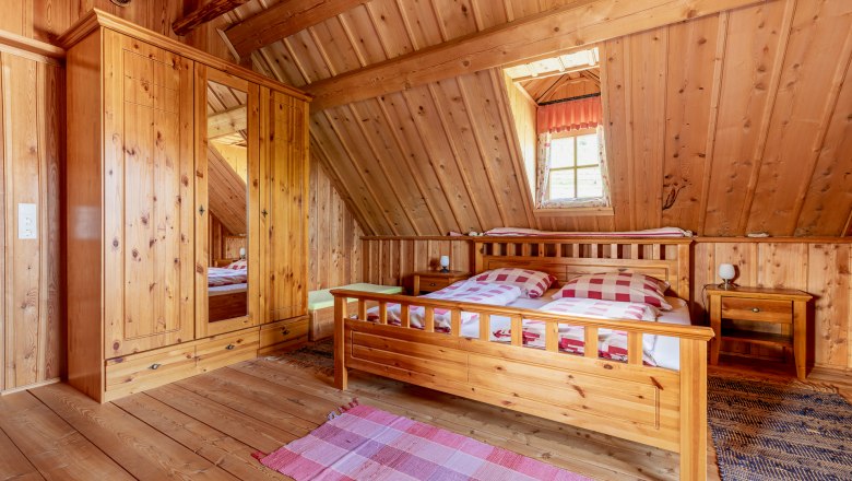 Schlafzimmer, © Wiener Alpen / Christian Kremsl Schlafzimmer mit Holzvertäfelung, großem Doppelbett, Holzkasten, Teppichen und einem Fenster.