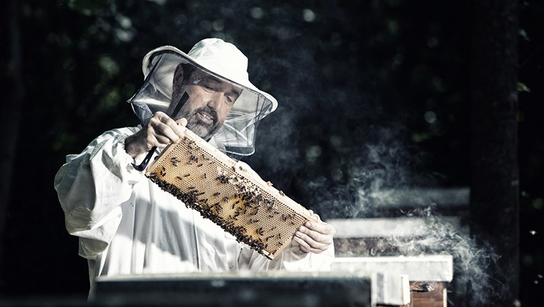Imkerei Gottfried Schwarz, © Imkerei Gottfried Schwarz Ein Imker im Schutzanzug hält einen Rahmen mit Bienenwaben und Bienen, umgeben von Rauch.