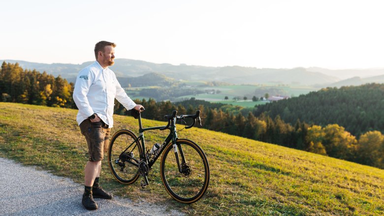 Wirt Andreas Ottner ist passionierter Biker, © Niederösterreich Werbung/Mara Hohla Ein Mann in weißem Hemd steht mit einem Fahrrad auf einem Hügel mit Blick auf eine grüne Landschaft.