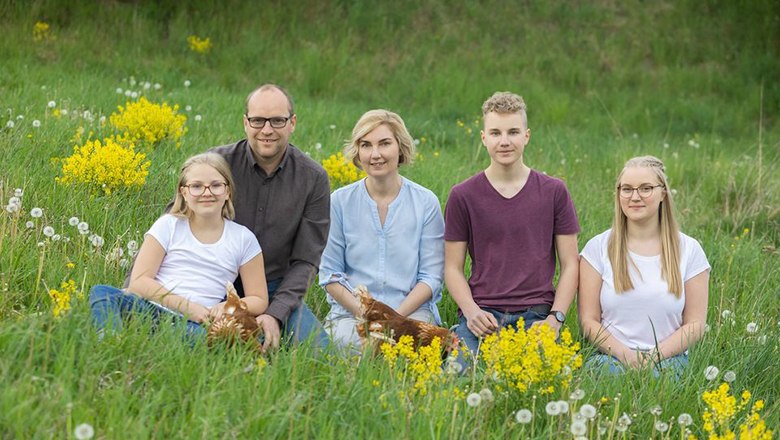 Familie Trimmel, © Biohof Trimmel Eine Familie mit drei Kindern sitzt mit zwei Hühnern auf einer grünen Wiese mit gelben Blumen.
