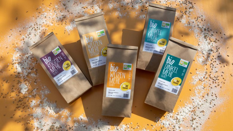 Bio-Mehle der Familie Trimmel, © Biohof Trimmel / Ulrike Korntheuer Fünf verschiedene Packungen Mehl auf orangem Hintergrund, umgeben von verstreutem Mehl und Körnern.