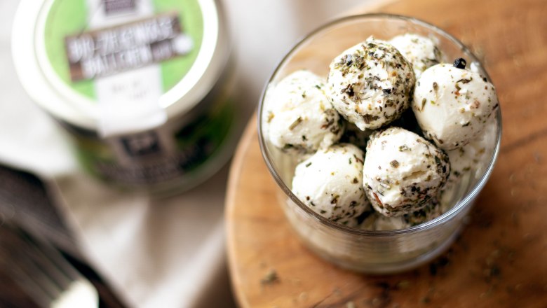 Ziegenkäsebällchen, © Sooo gut schmeckt die Bucklige Welt/Viktoria Kornfeld Ein Glas gefüllt mit Ziegenkäsebällchen, bestreut mit Kräutern, auf einem runden Holzbrett. Im Hintergrund ein weiteres Glas Ziegenkäsebällchen mit grünem Etikett.