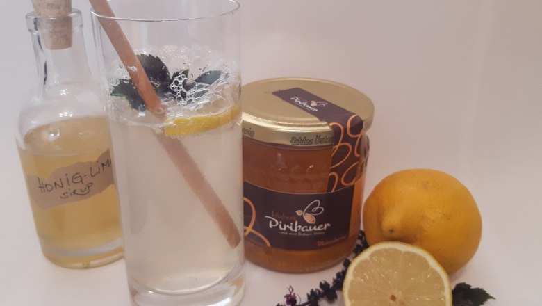 Honiglimonade, © Imkerei Piribauer Ein Glas Honiglimonade mit Minze und Zitrone, daneben Honigglas, Zitronen, Kräuter und Sirupflasche.