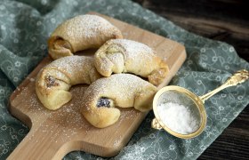 Vier Rahmkipferl auf einem Holzbrett, mit Puderzucker bestreut, daneben ein goldenes Sieb auf einem gemusterten Tuch., © Fam. Luckerbauer Vier Rahmkipferl auf einem Holzbrett mit Puderzucker bestreut, daneben ein Sieb mit Puderzucker.