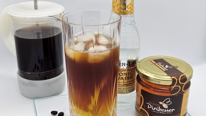 Cold-Brew-Tonic, © Imkerei Piribauer Ein Glas Cold-Brew-Tonic mit Eiswürfeln, daneben Honigglas, Glasflasche und Kaffeekanne auf Glasplatte.