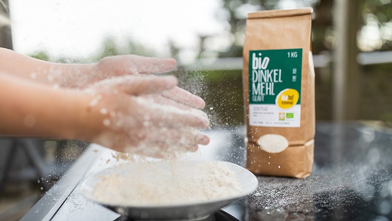 Biohof Trimmel, © Biohof Trimmel Hände klopfen Mehl über einem Teller, daneben eine Packung Bio-Dinkelmehl vom Biohof Trimmel.