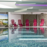 Hallenbad, © Vivea Gesundheitshotel Hallenbad mit roten Liegestühlen, Steinwänden und einem großen Wandbild.