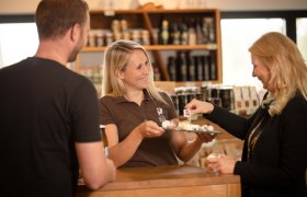 Hofladen, © AMA GENUSS REGION / Doris Schwarz König Drei Personen in einem Hofladen, eine lächelnde Frau bietet Käseproben an.