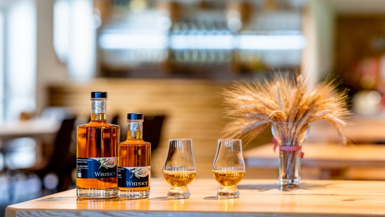 Whisky Kölbel, © Wiener Alpen, Christian Kremsl Zwei Whiskyflaschen und zwei Gläser mit Whisky stehen auf einem Holztisch, daneben ein Strauß Getreide.