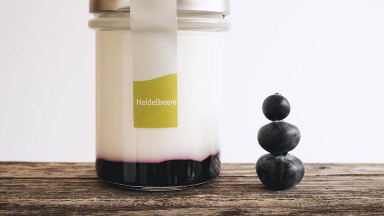 Joghurt Biohof am Stickelberg, © Anni Lechner Fotografie Ein Glas gefüllt mit Joghurt und Fruchtspiegel auf einem Holztisch, daneben 3 gestapelte Heidelbeeren.