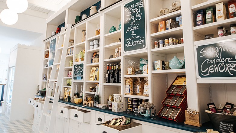 Eis-Greissler Hofladen innen, © Blochberger Eisproduktion GmbH Hofladen mit weißen Holzregalen und diversen Produkten wie Gewürzen, Schokoladen und Tees.