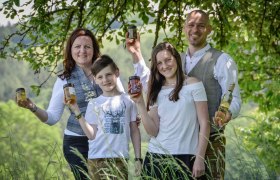 Eine Familie mit zwei Kindern steht auf einer grünen Wiese vor einem Baum und jede Person hält ein Glas Honig in der Hand.