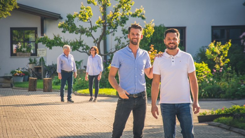 Zwei Männer stehen lächelnd im Vordergrund, zwei ältere Personen im Hintergrund, vor einem Haus mit Garten.