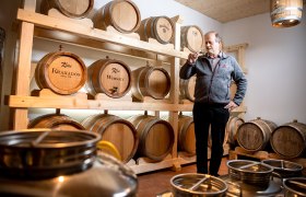 Ein Mann steht in einem Raum mit Holzfässern, die mit 'Kraxados' und 'Whisky' beschriftet sind, und hält ein Glas in der Hand.