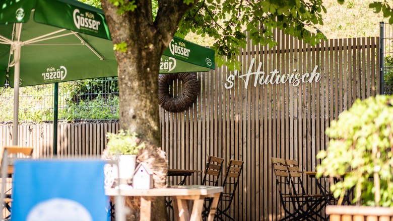 Ein Gastgarten mit Holztischen und -st&uuml;hlen, gr&uuml;nen Sonnenschirmen und einem Holzzaun mit der Aufschrift 's'Hutwisch'. in der Mitte steht ein Baum, der von einer Bank umgeben ist.