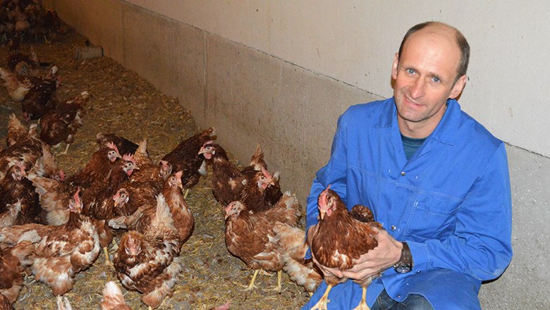 Ein Mann hält lächelnd ein Huhn in einem Hühnerstall, umgeben von vielen weiteren Hühnern.