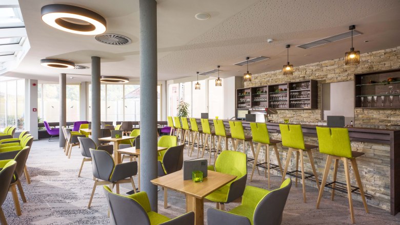 Modernes Café mit grünen und grauen Stühlen, Holztischen und einer langen Bar mit Steinwand.