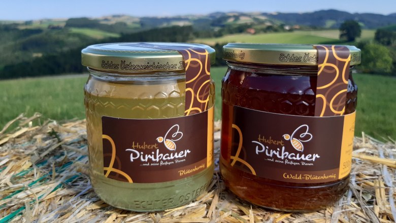 Ein Glas Blütenhonig und ein Glas Waldhonig auf Stroh vor einer grünen Landschaft.