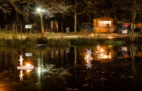 Ein See mit Lichtern und weihnachtlicher Dekoration, im Hintergrund Weg mit Adventmarktständen.