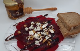 Rote Rüben Carpaccio mit Ziegenfrischkäse, Nüssen, Balsamico und Brot auf Glasteller, daneben Honig.