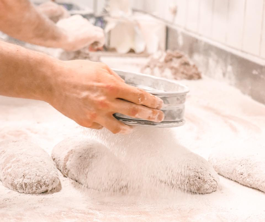 Hände sieben Mehl über Teigstücke auf einer bemehlten Arbeitsfläche in einer Bäckerei.