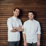 Zwei Männer in weißen Hemden stehen vor einer Holzwand und lächeln.