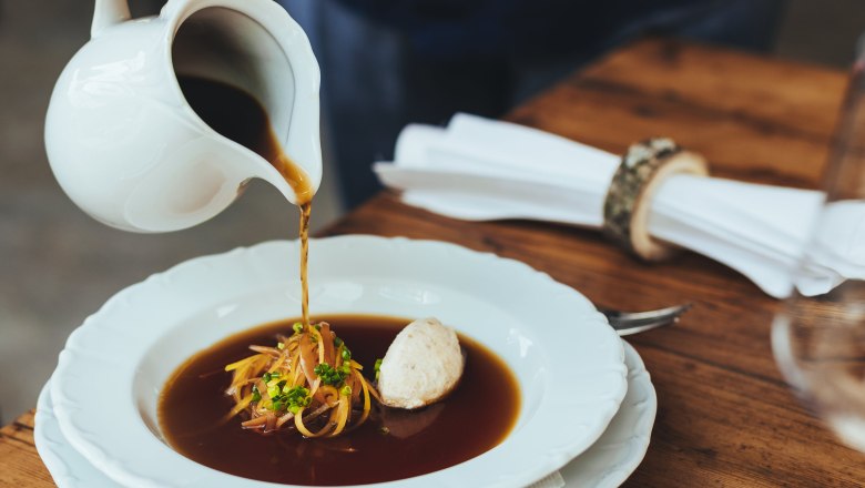 Ein Teller mit Suppe, Gemüse und Knödel, eine Hand gießt mit einer Kanne Suppe in das Teller.