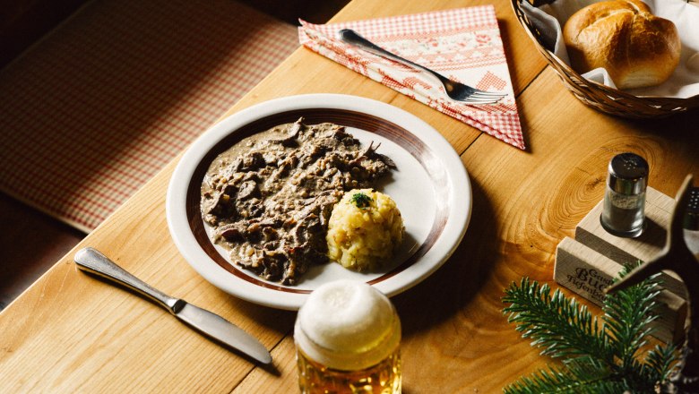 Teller mit Gericht, daneben Bierkrug, Serviette, Besteck, Brotkorb und Dekoration.