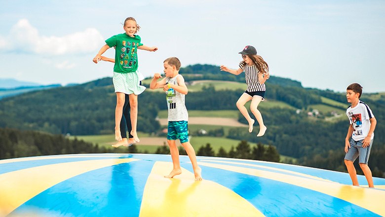 Vier Kinder springen auf einem bunten Hüpfpolster vor einer hügeligen Landschaft mit vielen Bäumen.