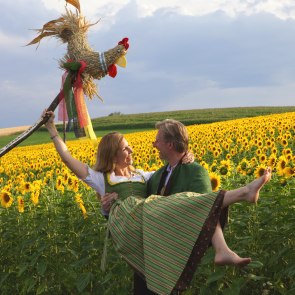 Mann trägt Frau im Arm, beide in Tracht, vor einem Sonnenblumenfeld.