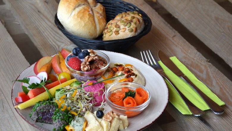 Bunter Frühstücksteller mit Hummus, Käse und Gemüse, daneben Besteck und ein Brotkorb.