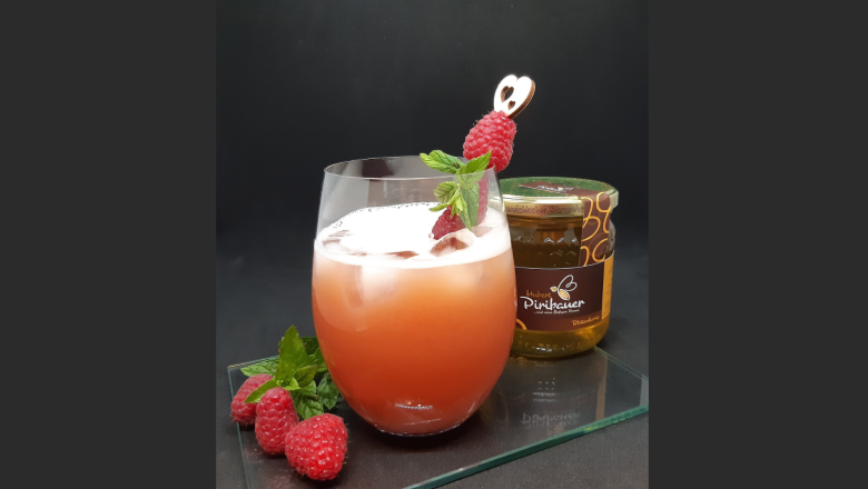 Ein Glas mit Honey Rider Cocktail, Eiswürfeln und Himbeerspieß, daneben Himbeeren und Honigglas.