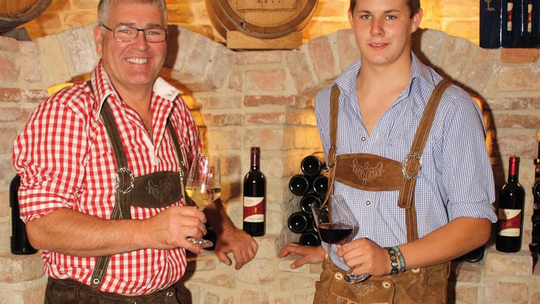 Zwei Männer in Tracht mit Weingläsern vor einer Ziegelwand.