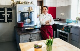 Eine Köchin in weißer Jacke und roter Schürze steht in einer Küche vor einem alten Herd.