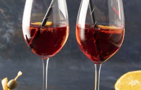 Zwei Weingläser mit Martini Vibrante Sprizz, Orangen und Strohhalmen, daneben Spieße mit Orangen und Oliven.