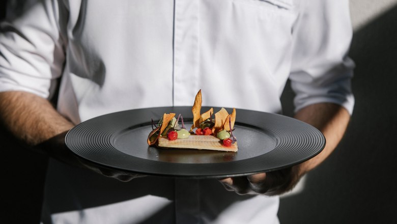 Person im weißen Hemd hält einen Teller mit einem kunstvoll angerichteten Gourmetgericht.