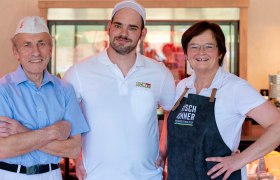 Drei lächelnde Personen in Arbeitskleidung stehen in einer Fleischerei.