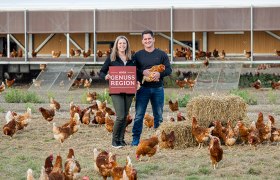 Eine Frau mit einem roten Schild der "AMA Genusregion" und ein Mann mit einem Huhn stehen vor einem Hühnerstall umgeben von vielen Hühnern.