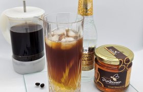 Ein Glas Cold-Brew-Tonic mit Eiswürfeln, daneben Honigglas, Glasflasche und Kaffeekanne auf Glasplatte.