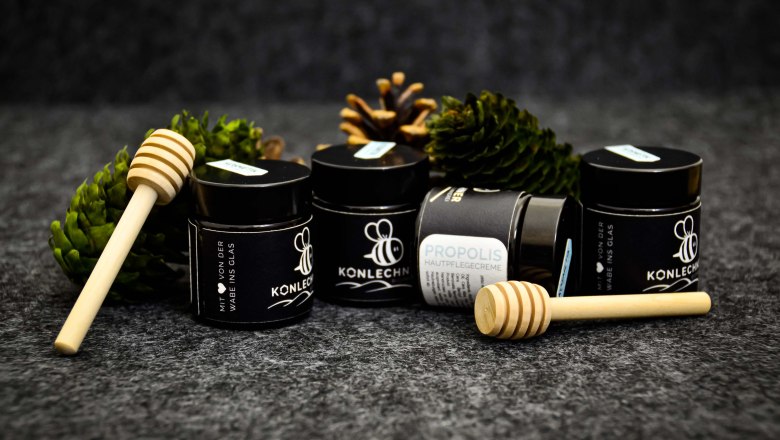 Vier kleine Gläser Propolis-Creme mit schwarzem Etikett mit Tannenzapfen und Honiglöffeln vor schwarzem Hintergrund., © Bernhard Singer